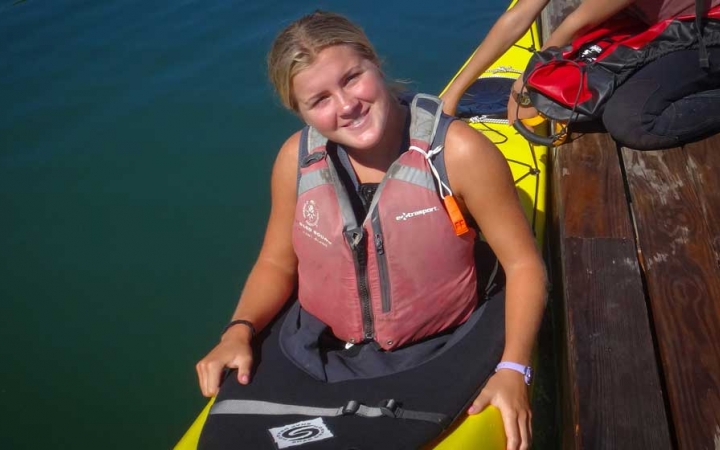 A young person wearing a life jacket sits in a kayak beside a dock and smiles at the camera